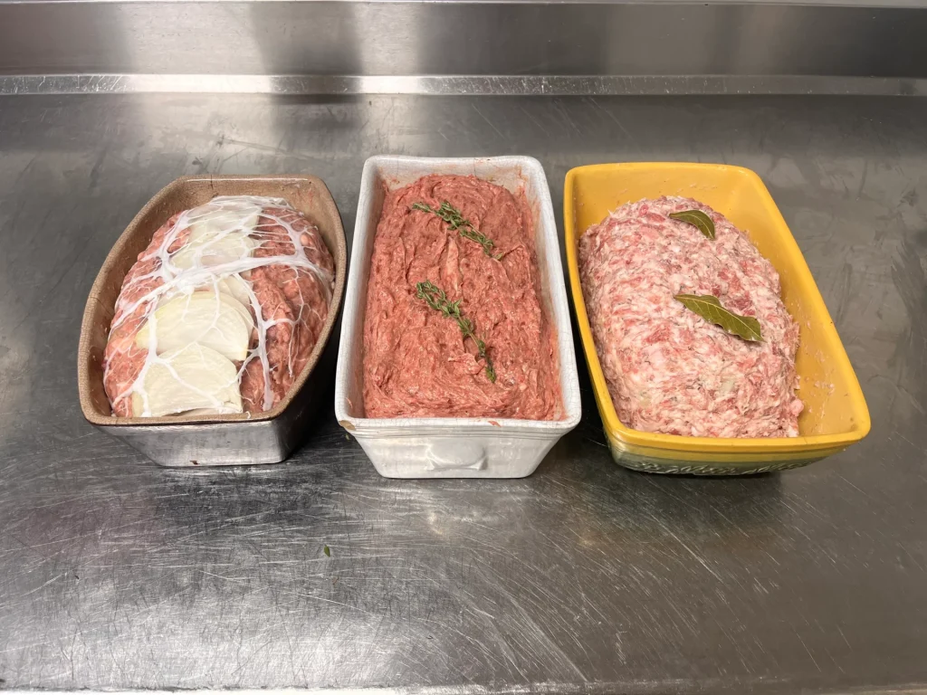 Trois plateaux sur une surface métallique contenant différents types de viande crue assaisonnée : le premier est du porc persillé, le deuxième est de la viande hachée aux herbes et le troisième est une viande hachée plus légère aux feuilles de laurier.