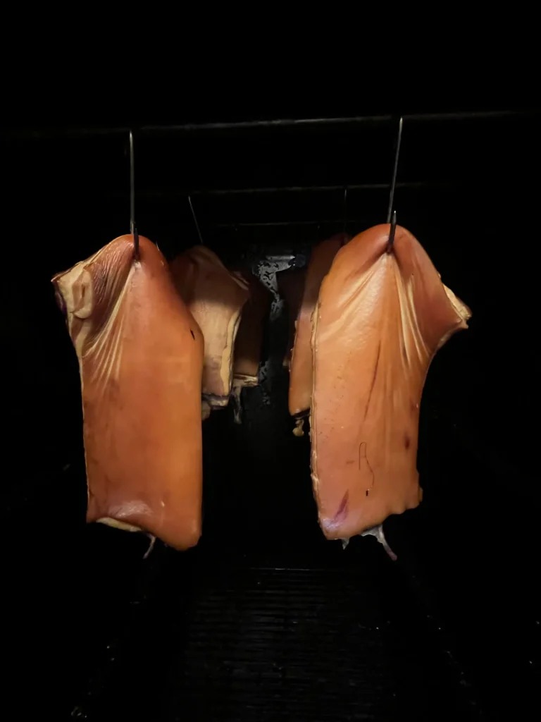 Des jambons entiers suspendus dans une chambre de fumage sombre, doucement éclairés par l'avant, mettant en valeur leurs surfaces brillantes et fumées.