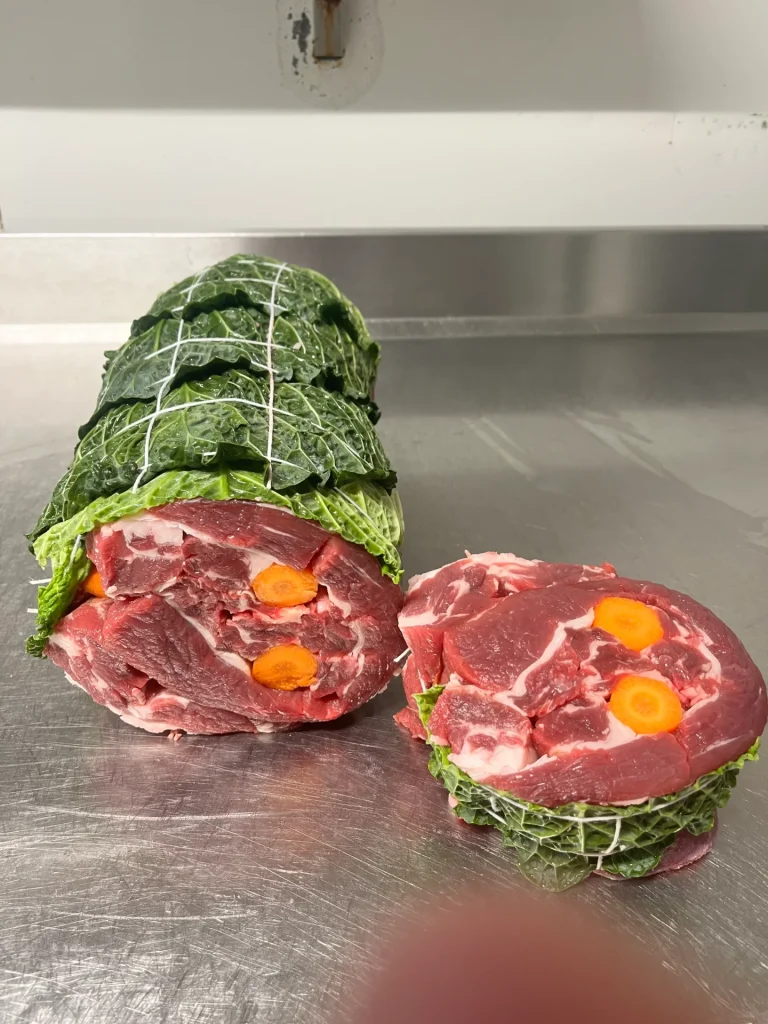 Un morceau roulé de viande crue enveloppé dans des feuilles de chou vert, attaché avec de la ficelle, à côté de deux tranches transversales montrant les couches intérieures avec des carottes, sur une surface en acier inoxydable.